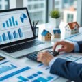A person working at a desk with financial documents, a laptop, a calculator, and small house models, analyzing real estate short sales.
