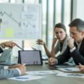 A group of real estate investors and professionals in a meeting room reviewing property charts and documents, discussing challenges related to short sales.