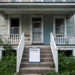 A rundown house with peeling paint, overgrown yard, and broken windows. A clipboard with budget numbers and renovation plans sits on the front steps