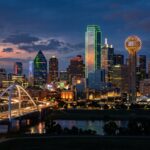 A detailed view of the Dallas-Fort Worth skyline, emphasizing the unique architectural profiles of the skyscrapers and the Reunion Tower. The scene features vibrant city lighting at dusk, with glowing windows and neon accents against a deepening sky, while maintaining the original composition and urban landscape.