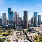 skyline of Downtown Houston, Texas, emphasizing the glass-clad skyscrapers, the distinct architectural silhouettes of the major high-rise buildings, and the urban density of the city center under a bright, clear sky.