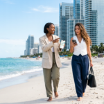 Two stylish Gen Z women in contemporary business-casual attire walk along the white sand of a Miami beach, gesturing toward a skyline of modern glass condominiums. They hold smartphones displaying real estate data and maps, their expressions focused as they engage in a professional discussion. The scene is illuminated by bright, natural sunlight, highlighting the turquoise water and the sleek architectural details of the nearby luxury towers.