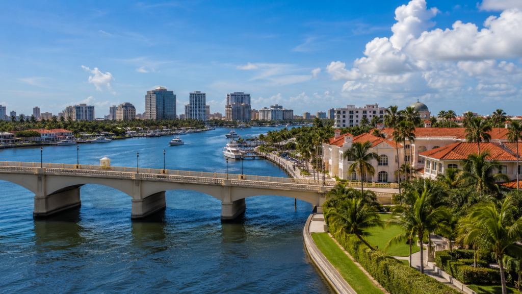 Aerial view of Palm Beach County waterfront with a bridge, luxury homes, palm trees, and downtown buildings along the Intracoastal Waterway.