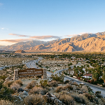 A scenic view of the Riverside–San Bernardino skyline nestled against the backdrop of the rugged, snow-capped San Bernardino Mountains. The landscape features a blend of modern and historic architecture, with vibrant palm trees and lush Mediterranean greenery lining the streets. Warm, golden hour sunlight casts a soft amber glow over the valley, highlighting the textures of the urban structures and the surrounding arid hills with cinematic lighting and clear atmospheric depth.