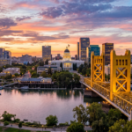 A vibrant urban landscape of Sacramento, California, featuring the distinctive architecture of the downtown skyline and the iconic gold-toned Tower Bridge spanning the Sacramento River. The scene is illuminated by the warm, directional glow of late afternoon sun, creating long shadows and shimmering reflections on the water's surface, while enhancing the textures of the city buildings and the surrounding leafy trees.