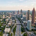 A wide-angle aerial view of the Atlanta metropolitan skyline, featuring iconic skyscrapers like the Bank of America Plaza and the cylindrical Westin Peachtree Plaza rising above the urban center. The dense city landscape is interwoven with the lush green tree canopies characteristic of the region, while the sprawling interstate highway system below is filled with the steady flow of daytime traffic. The scene is captured in the clear, natural light of a late afternoon, highlighting the varied architectural textures of glass, steel, and concrete against a soft, vast sky.