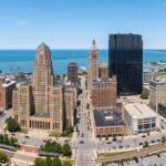 A wide-angle panoramic view of the Buffalo, New York skyline, highlighting the distinct architectural details of City Hall and the surrounding downtown buildings. The composition captures the urban layout with natural daylight, emphasizing the metallic and stone textures of the cityscape against a clear atmosphere, with the serene expanse of Lake Erie lining the horizon.