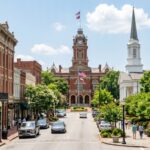 Transform the scene into a photorealistic view of Macon, Georgia, featuring its distinctive historic architecture and lush Southern greenery. Enhance the natural daylight to create a clear, scenic atmosphere with detailed textures on the buildings and vibrant foliage throughout the landscape.