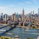 A wide panoramic view of the Manhattan skyline across the water, featuring prominent skyscrapers like the Empire State Building and One World Trade Center. The sprawling urban landscape stretches across the horizon under a clear sky, with the river in the foreground and the intricate steel structures of suspension bridges. Sunlight reflects off the glass and stone facades of the buildings, capturing the dense architectural variety and the iconic silhouette of the New York City waterfront from an elevated perspective.
