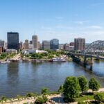 Scenic view of the Peoria skyline along the Illinois River, featuring the architectural details of the downtown buildings and the structural design of the Murray Baker Bridge. The landscape is captured with natural daylight, emphasizing the textures of the water's surface and the greenery of the riverfront. Clear atmospheric conditions provide depth and clarity to the urban scene while maintaining the original composition and city layout.