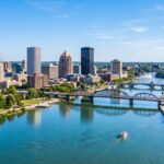 A sweeping panoramic view of the Rochester, New York skyline, emphasizing the architectural silhouettes and the flowing Genesee River in the foreground. Enhance the clarity of the bridges and waterfront buildings, with vibrant lighting reflecting off the water's surface. The scene should feel expansive and detailed, capturing the distinct urban layout and natural landmarks under a clear, bright sky.