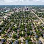 A distant aerial perspective of the Rockford, Illinois housing landscape, showcasing a vast expanse of residential neighborhoods with diverse architectural styles, tree-lined avenues, and a dense arrangement of suburban rooftops sprawling across the Midwestern terrain.