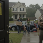 A uniformed sheriff with a badge stands by the front door of a suburban house, watching as a family carries cardboard boxes and personal belongings toward a parked car. A bright foreclosure notice is taped to the front door. The parents look somber and weary, carrying heavy containers of household items, while a young child clutches a single toy. The scene is set in a quiet residential neighborhood under a gray, overcast sky, capturing a heavy and somber atmosphere.