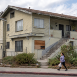A small apartment building showing some signs of distress as tenants walk by.