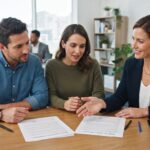 A professional female real estate investor presenting two different contractual agreements to a husband and wife in a bright, modern office setting. The investor is gesturing toward the documents on a table, explaining the nuances between lease-purchase and rent-to-own options. The scene emphasizes a collaborative atmosphere with natural lighting, focused expressions, and crisp details on the paperwork and professional attire.
