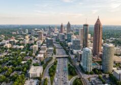 A wide-angle aerial view of the Atlanta metropolitan skyline, featuring iconic skyscrapers like the Bank of America Plaza and the cylindrical Westin Peachtree Plaza rising above the urban center. The dense city landscape is interwoven with the lush green tree canopies characteristic of the region, while the sprawling interstate highway system below is filled with the steady flow of daytime traffic. The scene is captured in the clear, natural light of a late afternoon, highlighting the varied architectural textures of glass, steel, and concrete against a soft, vast sky.