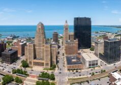 A wide-angle panoramic view of the Buffalo, New York skyline, highlighting the distinct architectural details of City Hall and the surrounding downtown buildings. The composition captures the urban layout with natural daylight, emphasizing the metallic and stone textures of the cityscape against a clear atmosphere, with the serene expanse of Lake Erie lining the horizon.