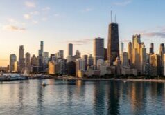 Photorealistic Chicago skyline with detailed architectural textures on the skyscrapers and glass facades. The scene is set during a clear golden hour, with warm, directional sunlight casting soft shadows and highlighting the city's silhouette. The surrounding water features natural ripples and clear reflections of the buildings and the gradient of the sky.
