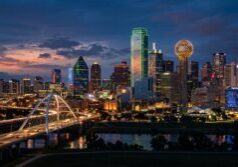 A detailed view of the Dallas-Fort Worth skyline, emphasizing the unique architectural profiles of the skyscrapers and the Reunion Tower. The scene features vibrant city lighting at dusk, with glowing windows and neon accents against a deepening sky, while maintaining the original composition and urban landscape.