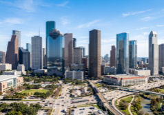 skyline of Downtown Houston, Texas, emphasizing the glass-clad skyscrapers, the distinct architectural silhouettes of the major high-rise buildings, and the urban density of the city center under a bright, clear sky.