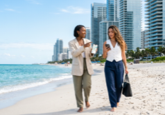 Two stylish Gen Z women in contemporary business-casual attire walk along the white sand of a Miami beach, gesturing toward a skyline of modern glass condominiums. They hold smartphones displaying real estate data and maps, their expressions focused as they engage in a professional discussion. The scene is illuminated by bright, natural sunlight, highlighting the turquoise water and the sleek architectural details of the nearby luxury towers.