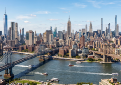 A wide panoramic view of the Manhattan skyline across the water, featuring prominent skyscrapers like the Empire State Building and One World Trade Center. The sprawling urban landscape stretches across the horizon under a clear sky, with the river in the foreground and the intricate steel structures of suspension bridges. Sunlight reflects off the glass and stone facades of the buildings, capturing the dense architectural variety and the iconic silhouette of the New York City waterfront from an elevated perspective.