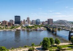 Scenic view of the Peoria skyline along the Illinois River, featuring the architectural details of the downtown buildings and the structural design of the Murray Baker Bridge. The landscape is captured with natural daylight, emphasizing the textures of the water's surface and the greenery of the riverfront. Clear atmospheric conditions provide depth and clarity to the urban scene while maintaining the original composition and city layout.