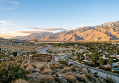 A scenic view of the Riverside–San Bernardino skyline nestled against the backdrop of the rugged, snow-capped San Bernardino Mountains. The landscape features a blend of modern and historic architecture, with vibrant palm trees and lush Mediterranean greenery lining the streets. Warm, golden hour sunlight casts a soft amber glow over the valley, highlighting the textures of the urban structures and the surrounding arid hills with cinematic lighting and clear atmospheric depth.