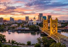 A vibrant urban landscape of Sacramento, California, featuring the distinctive architecture of the downtown skyline and the iconic gold-toned Tower Bridge spanning the Sacramento River. The scene is illuminated by the warm, directional glow of late afternoon sun, creating long shadows and shimmering reflections on the water's surface, while enhancing the textures of the city buildings and the surrounding leafy trees.