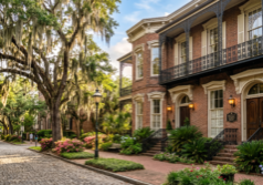 Reimagine the scene with characteristic Savannah, Georgia architectural elements, featuring historic brickwork, ornate wrought iron railings, and grand windows. Incorporate flowing Spanish moss draped from the surrounding trees and lush, manicured southern greenery. Apply a soft, warm daylight to highlight the textures of the facades, creating an inviting and professional real estate aesthetic that captures the charm of the historic district.