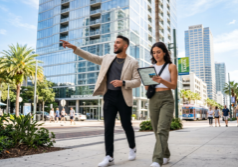 A stylish Gen Z male real estate investor and his girlfriend walking through the sun-drenched streets of downtown Tampa. The man, dressed in contemporary smart-casual attire, points toward a modern glass high-rise building while his girlfriend observes a digital tablet showing property details. The surrounding urban landscape features palm-lined sidewalks, sleek coastal architecture, and the bright, clear light of a Florida afternoon reflecting off the glass facades of nearby skyscrapers.