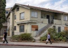 A small apartment building showing some signs of distress as tenants walk by.