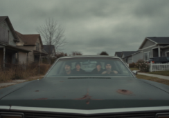A group of suspicious individuals in a weathered, nondescript car slowly cruising through a quiet suburban street. The neighborhood shows some homes with signs of neglect, with several houses featuring boarded-up windows and overgrown lawns. Other homes are in pristine condition. The scene is captured with a gritty, cinematic atmosphere under heavy, overcast skies even though it is day time, emphasizing a sense of tension as the occupants scan the properties for signs of abandonment while avoiding attracting attention from the neighbors.
