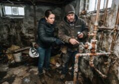 A female real estate investor in practical, professional attire and a male handyman in rugged workwear with the logo ForeclosureFlips.com inspect exposed copper plumbing and rusted utility shutoff valves in a basement. The setting is a cold, winterized foreclosure with visible water damage stains on the walls and frost clinging to the metal fixtures. Dusty, cool light filters through small, grimy windows, emphasizing the gritty textures of the derelict interior, damp surfaces, and scattered debris on the floor.